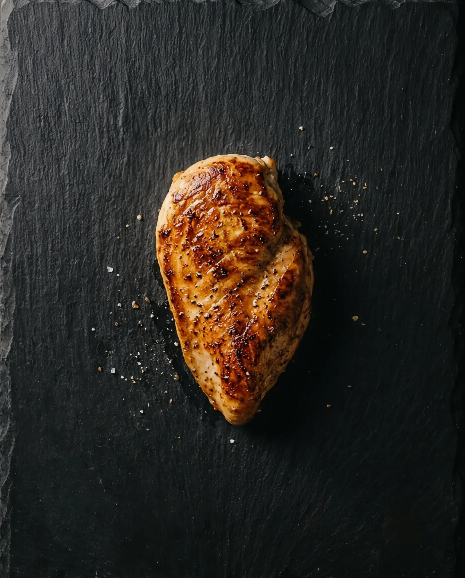A whole seared chicken fillet seen from above on dark slate